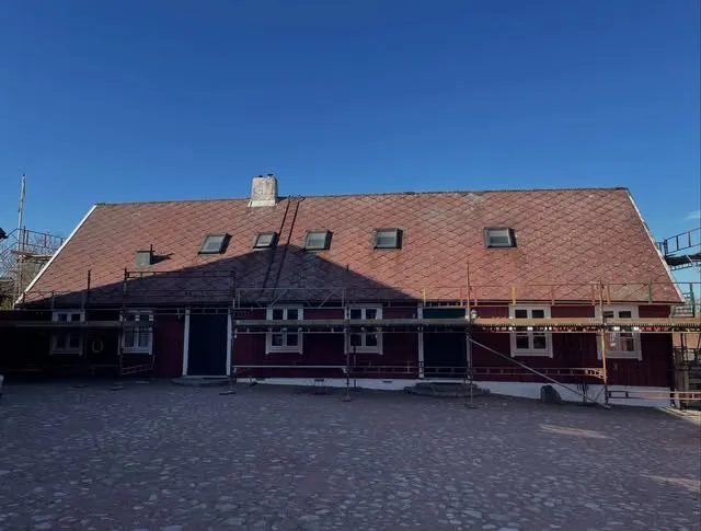 Rött gårdshus med rutmönstrat tak och byggnadsställning under blå himmel